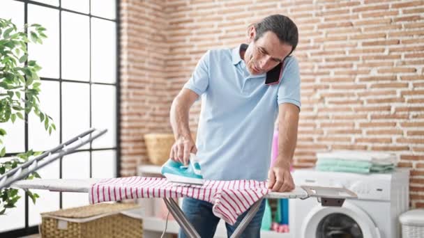 中年男性 洗濯室でスマートフォンアイロン掛け服で話す — ストック動画