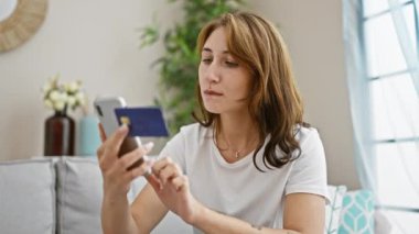 Akıllı telefon ve kredi kartıyla alışveriş yapan genç bir kadın evdeki kanepede oturuyor.