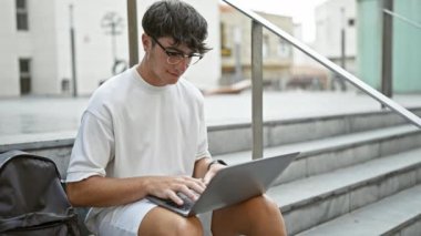Havalı genç İspanyol üniversiteli çocuk, kendinden emin bir şekilde gözlüğünü çıkarıyor, kampüs merdivenlerinde oturuyor, dizüstü bilgisayarla internette gezinirken gün ışığını yakalıyor, gülümsüyor..