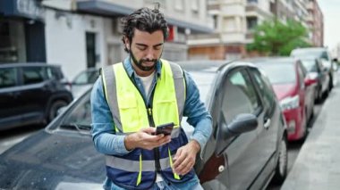Yansıtıcı yelek giymiş genç bir İspanyol sokakta akıllı telefon kullanıyor.