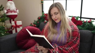 Evdeki Noel ağacının yanında kitap okuyan genç sarışın kadın.