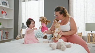 Kendine güvenen anne ve neşeli kız oyuncaklarla oyun oynuyorlar, güneşli bir yatak odasında rahat bir yatakta oturuyorlar, sıcak bir gülümsemeyle neşe ve sevgiyi ifade ediyorlar.