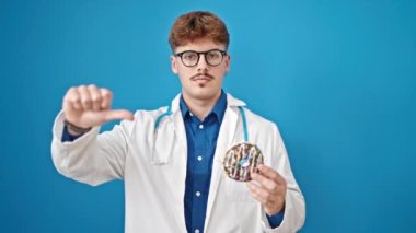 Genç İspanyol doktor elinde donut tutarak izole edilmiş mavi arka planda baş parmak hareketi yapıyor.