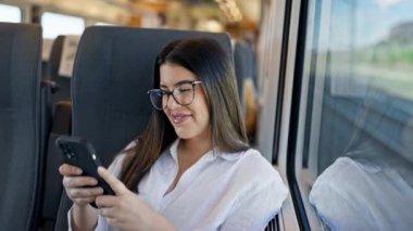 Genç ve güzel İspanyol kadın, tren istasyonunda oturmuş akıllı telefon kullanarak gülümsüyor.
