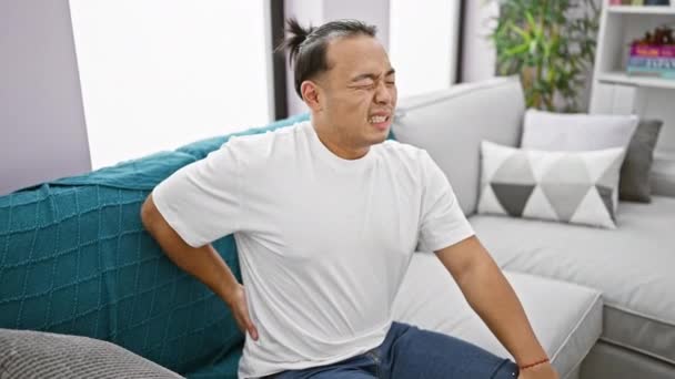 心配した若い中国人男性 深刻な腰痛に苦しんで 自宅のソファーに不幸に座って脊髄損傷の厳しい屋内現実を示しています — ストック動画