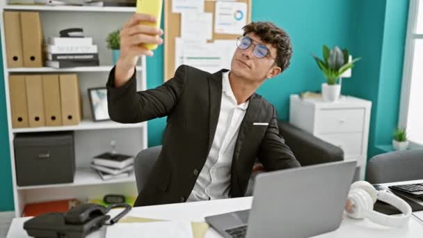 年轻英俊的上班族在办公室里惊慌失措 一边用手机发号施令 一边在笔记本电脑上拍自拍 — 图库视频影像