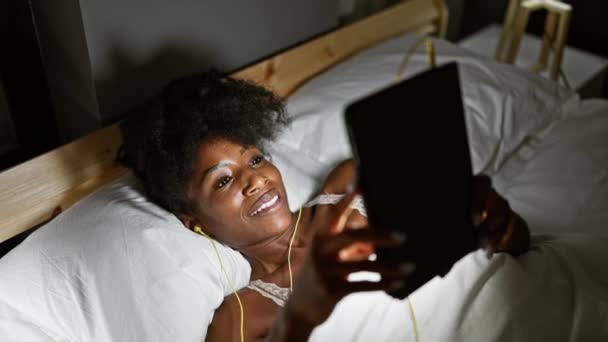 アフリカ系アメリカ人女性 タッチパッドとイヤホンを使用して寝室で笑顔ベッドに横たわる — ストック動画