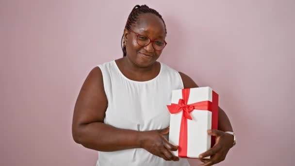 自信的非洲裔美国女人 戴着辫子和眼镜 漫不经心地靠着一面孤立的粉红墙壁 一边微笑着 一边竖起大拇指 手里拿着带着丝带的生日礼物 — 图库视频影像