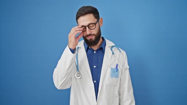 若いヒスパニックの男性医師は 孤立した青い背景の上にメガネを取る自信を持って微笑みます — ストック動画