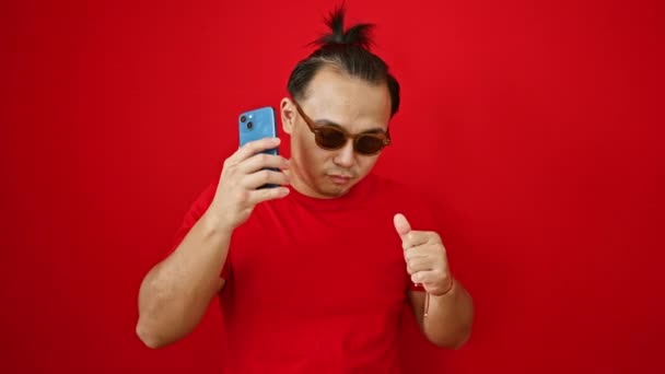 ファンキーなブタテールヘアスタイルの陽気な若いアジア人男性は 単離された赤い背景の上に喜んで踊り 携帯電話を保持し 彼のスマートフォンから音楽ストリーミングに完全に魅了 — ストック動画