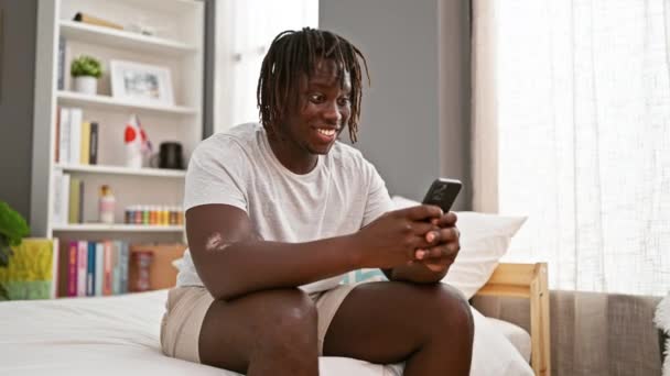 非洲裔美国人坐在床上用智能手机庆祝在卧室里 — 图库视频影像