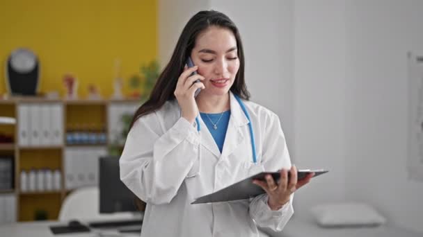 Young Beautiful Hispanic Woman Doctor Reading Document Clipboard Talking Smartphone — Stock Video