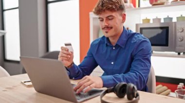 Genç İspanyol adam yemek odasında dizüstü bilgisayar kullanarak kredi kartını öpüyor.