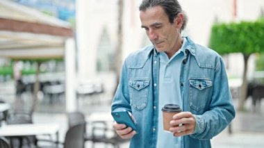 Orta yaşlı bir adam akıllı telefon kullanıyor kahve içiyor kahve dükkanının terasında gülümsüyor.