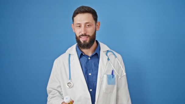 孤立した青い背景の上に丸薬瓶を保持している若いヒスパニック男性の医師 — ストック動画