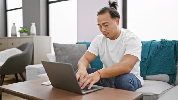陽気な若い中国人男性は屋内でリラックスし 彼のリビングルームのインテリアに座って腕を交差し 喜んで自宅のラップトップ上の技術を使用して — ストック動画