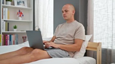 Yatak odasında dizüstü bilgisayar kullanan İspanyol bir adam stresli bir şekilde oturuyor.