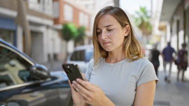 Neşeli genç sarışın kadın şehir sokaklarında akıllı telefonuyla eğlenceli bir selfie çekiyor. Şehir hayatının tadını çıkarırken gün ışığında güneşleniyor..