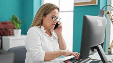 Orta yaşlı İspanyol iş kadını. Ofiste bilgisayar kullanıyor. Akıllı telefondan konuşuyor.