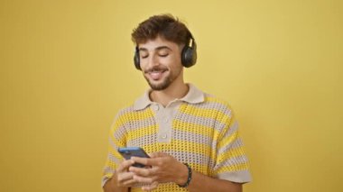 Havalı Arap adam güvenli bir şekilde telefonuyla neşeli bir müzik dinliyor ve dans ediyor. Mutluluk ve pozitifliğin izole edilmiş sarı bir arka planını yaratıyor.