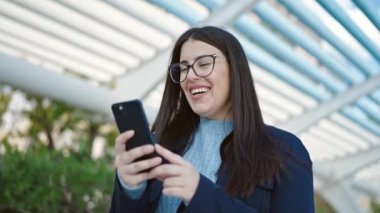 Genç İspanyol kadın parkta akıllı telefon kullanarak gülümsüyor.