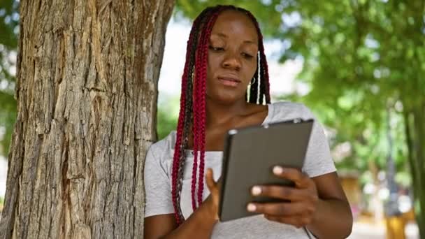 自信的非洲裔美国女人 美丽而快乐 靠在公园的树上 沐浴在阳光下 在户外使用她的触摸板时 闪烁着清凉而快乐的微笑 — 图库视频影像