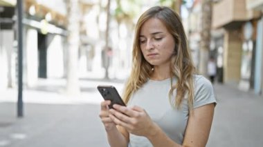 Neşeli genç sarışın kadın mutlu bir şekilde akıllı telefonuna dokunuyor güneşli bir caddede dijital bir sohbete dalıyor.