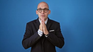 Ciddi yüzlü İspanyol bir iş adamı, izole edilmiş mavi arka planda dua jesti yapıyor.