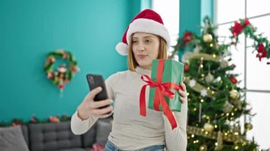 Elinde Noel hediyesi olan genç sarışın kadın akıllı telefonuyla selfie çekiyor.