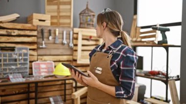 Yoğun bir marangozluk atölyesinde, dokunmatik dokunmatik bölmesine gülümseyen kendine güvenen genç sarışın kadın marangoz.