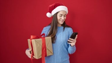 Kafkasyalı genç bir kadın yılbaşı şapkası takıyor. Akıllı telefon hediyesiyle izole edilmiş kırmızı arka planda.
