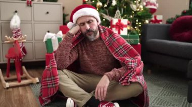 Genç kel adam Noel ağacının yanında oturmuş üzgün üzgün bakıyor.