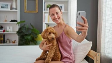 Köpeği gülümseyen beyaz bir kadın yatak odasındaki yatakta akıllı telefonuyla selfie çekiyor.
