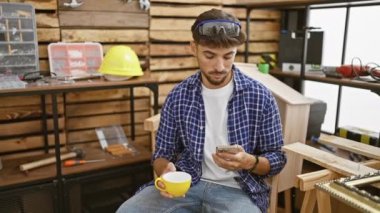 Yakışıklı genç bir Arap adam, mesleği marangoz, canlı marangozluk atölyesinde akıllı telefonuyla mesajlaşırken kahvesinin tadını çıkarıyor.