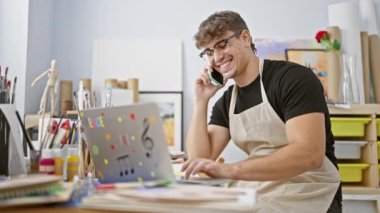Yakışıklı genç İspanyol ressam, akıllı telefonuyla derin sohbetler içinde, resim stüdyosunda dizüstü bilgisayarını ustalıkla kullanıyor. Etrafı boya fırçaları ve tuvallerle çevrili..