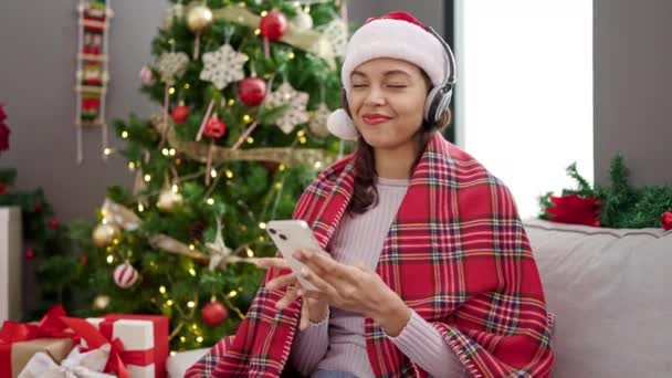 自宅でクリスマスの装飾によって音楽を聞く電話を使用して若い美しいヒスパニックの女性 — ストック動画