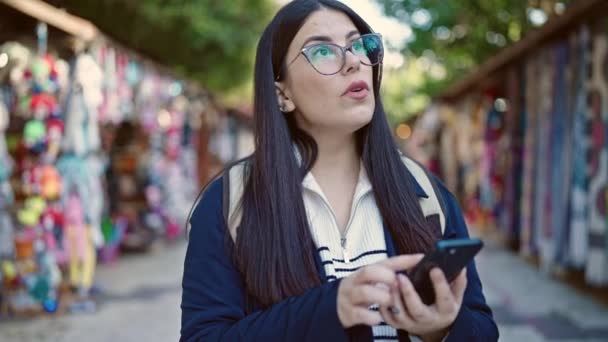 ストリートマーケットでスマートフォンを使用してバックパックを着用する若いヒスパニック女性観光客 — ストック動画
