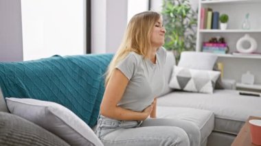 Çekici sarışın kadın ciddi karın ağrısı çekiyor. Kanepesinde uzanıyor. Kapalı bir yerde güneş ışığı altında, rahatsız bir şekilde dinleniyor..