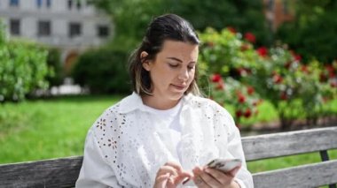 Güzel, genç, İspanyol bir kadın mutlu bir şekilde gülümsüyor. Parktaki bankta oturmuş akıllı telefon kullanıyor.
