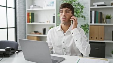 Zeki, genç İspanyol bir adam akıllı telefondan konuşuyor, ofis bilgisayarında çok düşünüyor, işyerindeki başarıyı somutlaştırıyor.