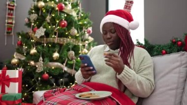 Saçları örgülü Afrikalı bir kadın akıllı telefon kullanıyor. Yılbaşı ağacının yanında oturuyor.