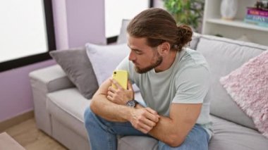 Genç İspanyol adam akıllı telefon kullanıyor. Kanepede oturuyor ve evde oturup düşünüyor.