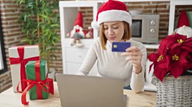 Laptop ve kredi kartıyla alışveriş yapan genç sarışın kadın Noel 'i yemek odasında kutluyor.
