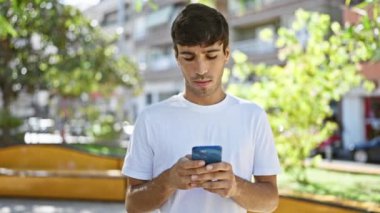 Havalı görünümlü İspanyol bir genç adam, ciddi bir ifadeyle, parkta akıllı telefonuyla mesajlaşmaya dalmış, açık havada güneşli bir havada, çekici, rahat bir yaşam tarzı yayıyor..