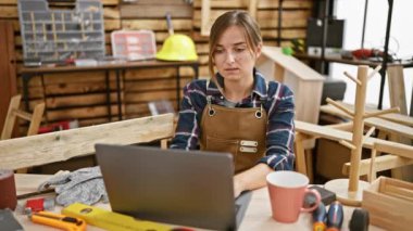 Gülümseyen genç sarışın marangoz, kollarını kavuşturmuş, marangozluk işini keyifle yürütüyor. İç mekandaki atölyede talaş ve kereste arasında.