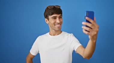 Havalı görünümlü genç İspanyol gözlüklü adam, kendinden emin bir şekilde neşeli bir selfie çekiyor, mavi duvarın karşısında tek başına duruyor..