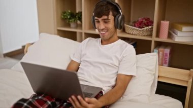 Uyanık ve gülümseyen, kendine güvenen genç İspanyol bir adam pijamalarıyla, laptopuyla yatakta oturup, yatak odasındaki kulaklıkla müzik dinlemekten zevk alır.