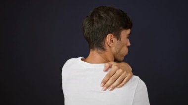 Yoğun sırt ağrısı çeken sıkıntılı genç bir İspanyol adam, siyah bir arka planda izole edilmiş, konsantre ve ciddi bir ifadeyle başka tarafa bakarken bir yaraya dokunuyor.