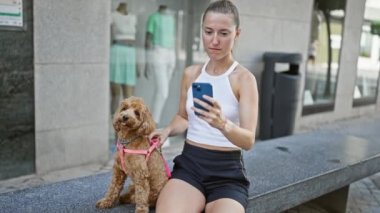 Köpekli beyaz bir kadın akıllı telefonuyla fotoğraf çekiyor. Sokakta bankta oturuyor.