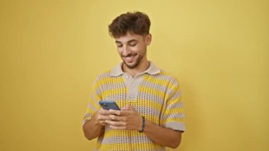Yakışıklı genç Arap adam, akıllı telefonuyla kendinden emin bir şekilde mesaj atıyor, neşeli bir şekilde izole edilmiş sarı arka plana gülümsüyor.